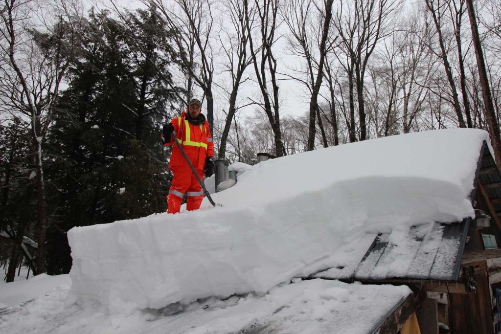 a man shovelling many feet of snow off the roof of a rustic house